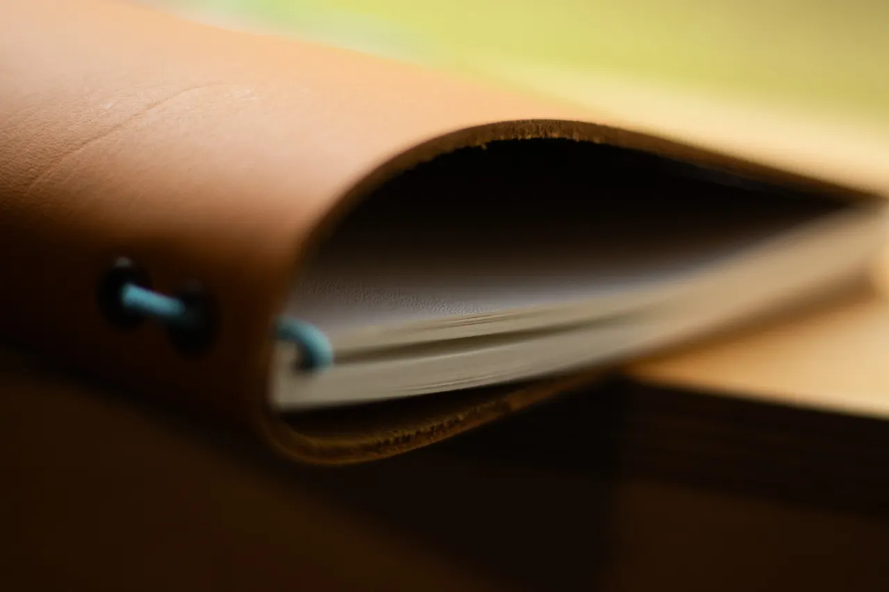 A close-up of a tan leather journal cover curling open, the pages and its blue binding cord just visible inside