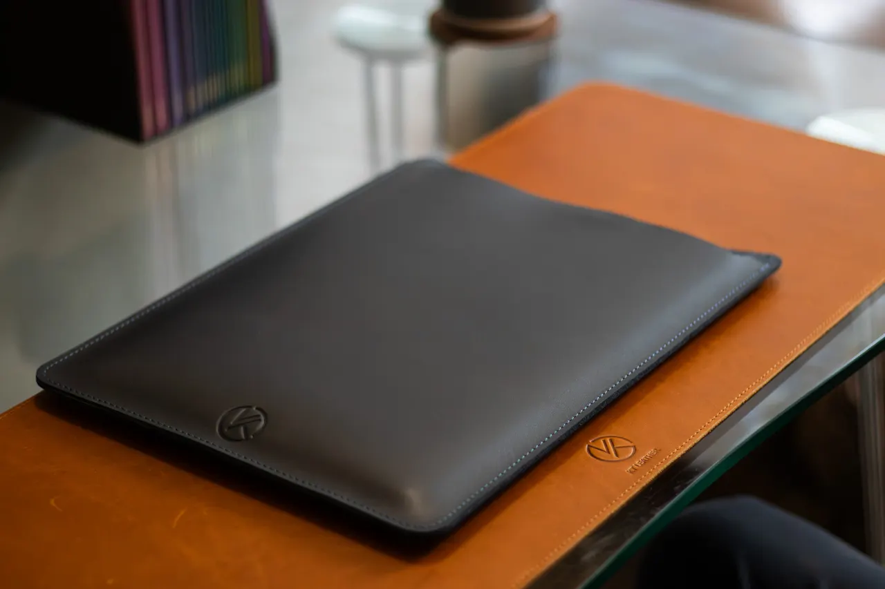 A charcoal leather laptop sleeve with the embossed KT mark, lying on a tan leather desk mat on a glass table