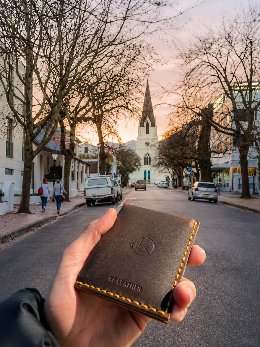 A dark brown KT Leather wallet with yellow stitching held up on Dorp Street in Stellenbosch, the white spire of the Moederkerk and a pink sunset behind it