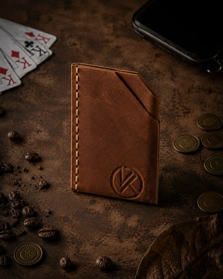 The Duo card wallet in tan leather standing on a dark weathered bench, the embossed KT mark facing out, brass coins and playing cards around it