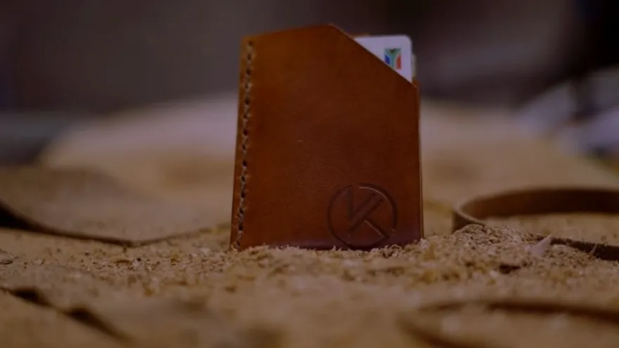 The Duo card wallet in tan leather standing upright in sawdust, an ID card showing above the embossed KT mark