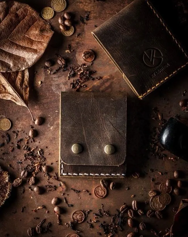 The Monty wallet in dark brown leather with brass press studs, on a rusted surface scattered with coffee beans and coins