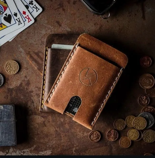 The Slyde wallet in tan leather, a card sliding out of the thumb slot, coins and playing cards on a worn steel bench